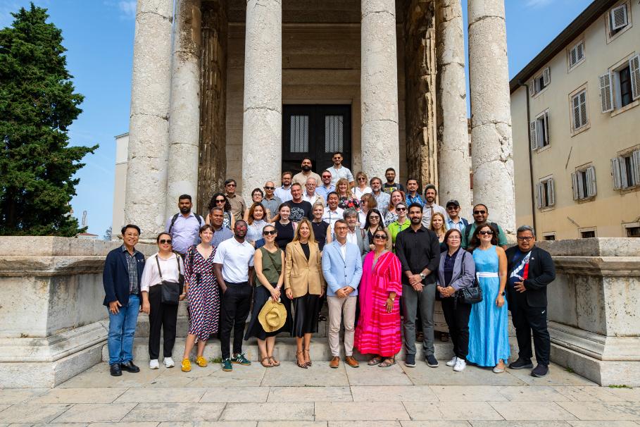 Group of professionals wearing various colours, smiling, standing on tiered steps going up to large columns in front of a hall.
